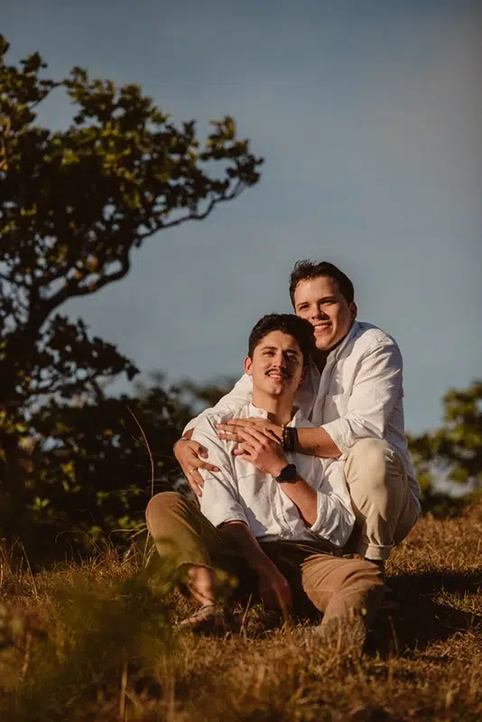 Love & Serenity: A Sunset Moment for an LGBTQ+ Couple A gay couple sitting together, embracing as they watch the sunset, with a blue sky in the background.
