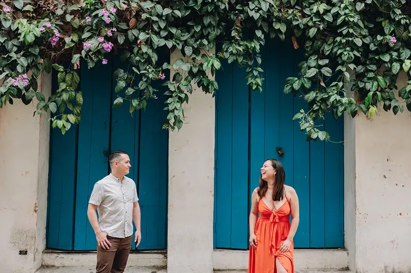 Joice & Diego: The faces behind the lens Joice and Diego, destination wedding photographers, smiling in front of a colorful wall in Cartagena