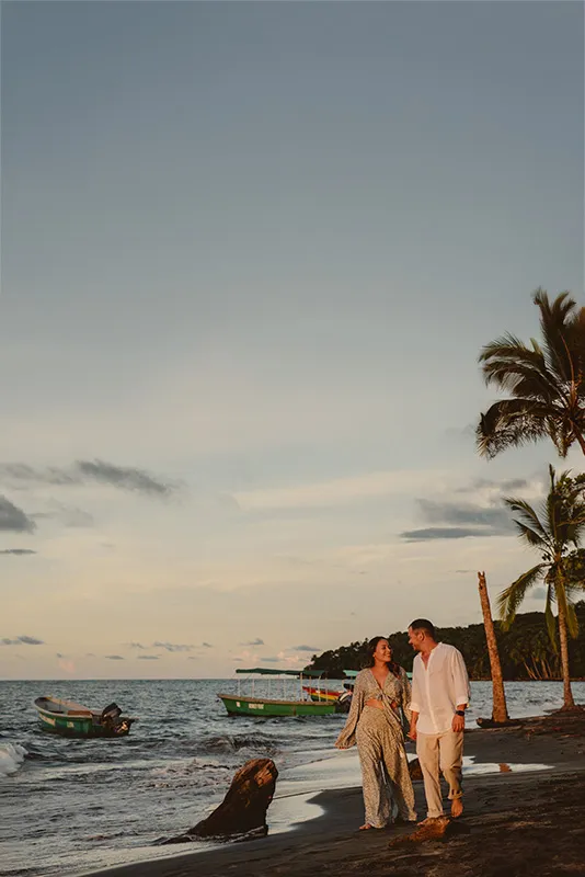 Couple walking on the beach with sunset light on their faces and fishing boats behind them.