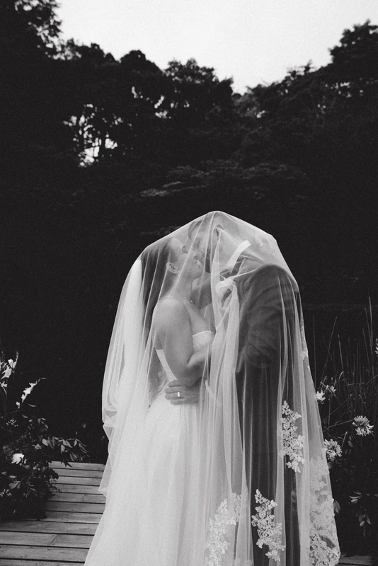 Wedding Photography in Costa Rica: bride veiled in a sheer wedding veil, standing before a dark tropical forest in a black and white portrait