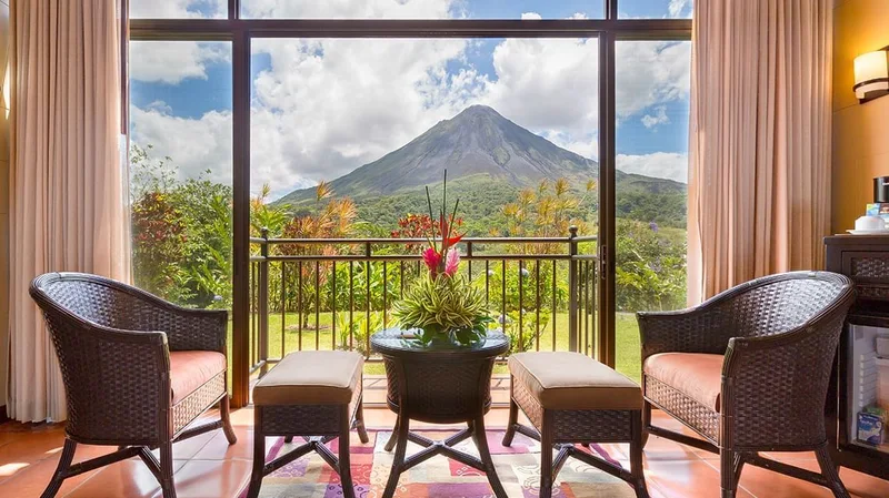 Hotel room with wicker chairs and a balcony framing Arenal Volcano at Arenal Kioro Suites & Spa, La Fortuna, Costa Rica