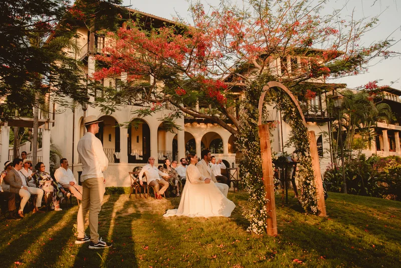 Bride and groom at a wooden ceremony arch under a red flame tree with guests and Spanish-colonial buildings behind them in Las Catalinas, Costa Rica