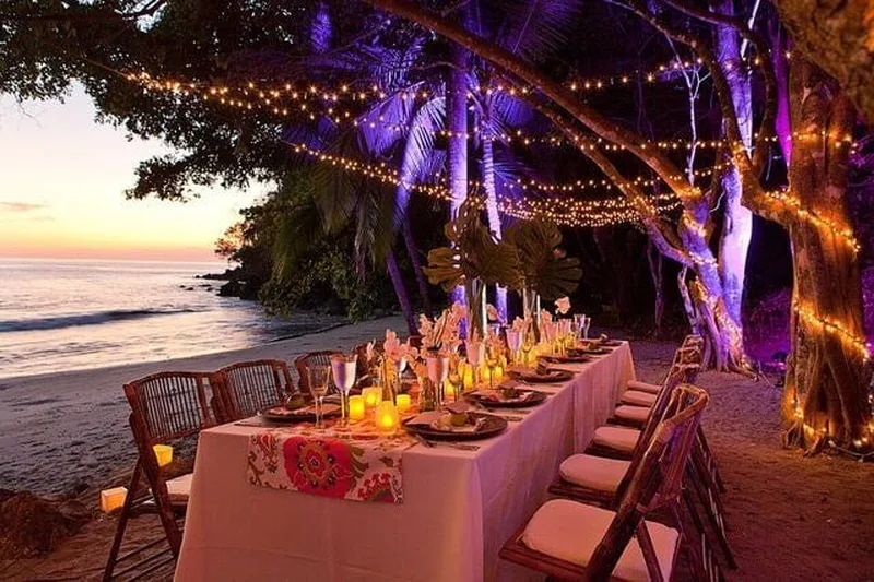 Long candlelit wedding dinner table on the beach under a tree with string lights at sunset at Tulemar Resort, Manuel Antonio