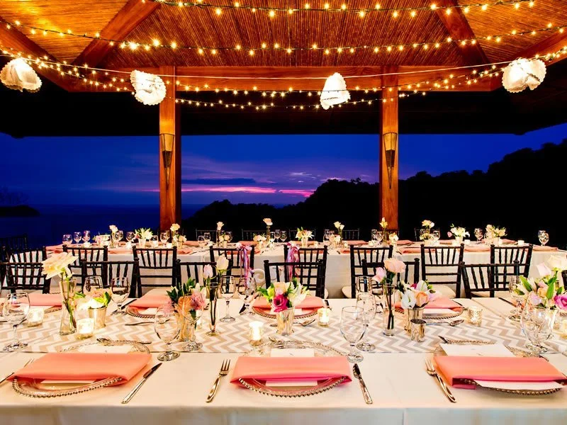 Wedding reception tables with pink napkins under a string-lit wooden pavilion at dusk at Villa Punto de Vista, Manuel Antonio, Costa Rica